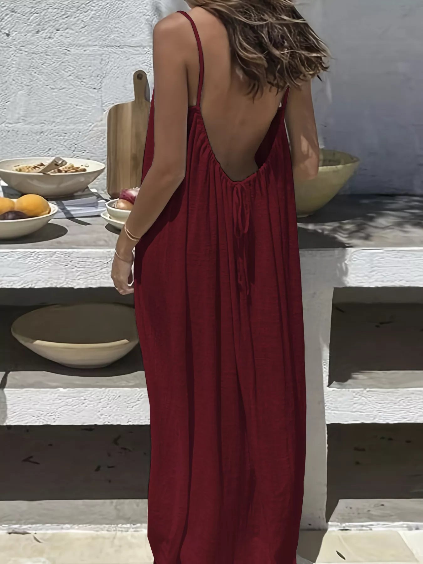 Woman in sleeveless red backless maxi dress outdoors near a table with fruits and bowls