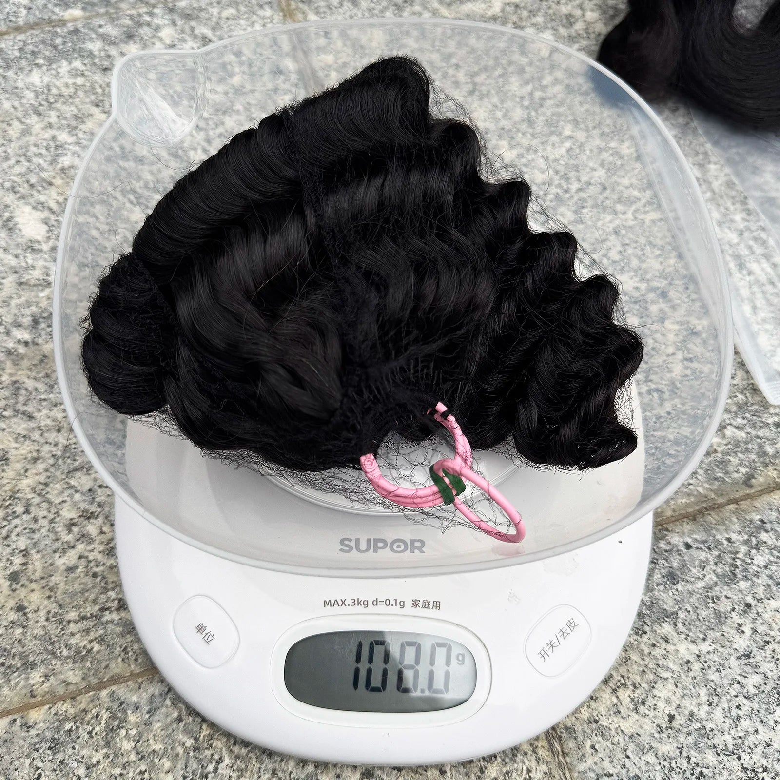 Curly black hair bundle on digital scale showing 108.9 grams, hair accessory visible